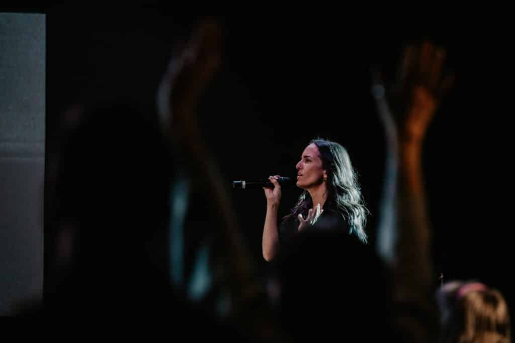 A woman singing into a microphone while performing on stage, with blurred hands raised in the foreground. The background features a dark setting that highlights the singer.