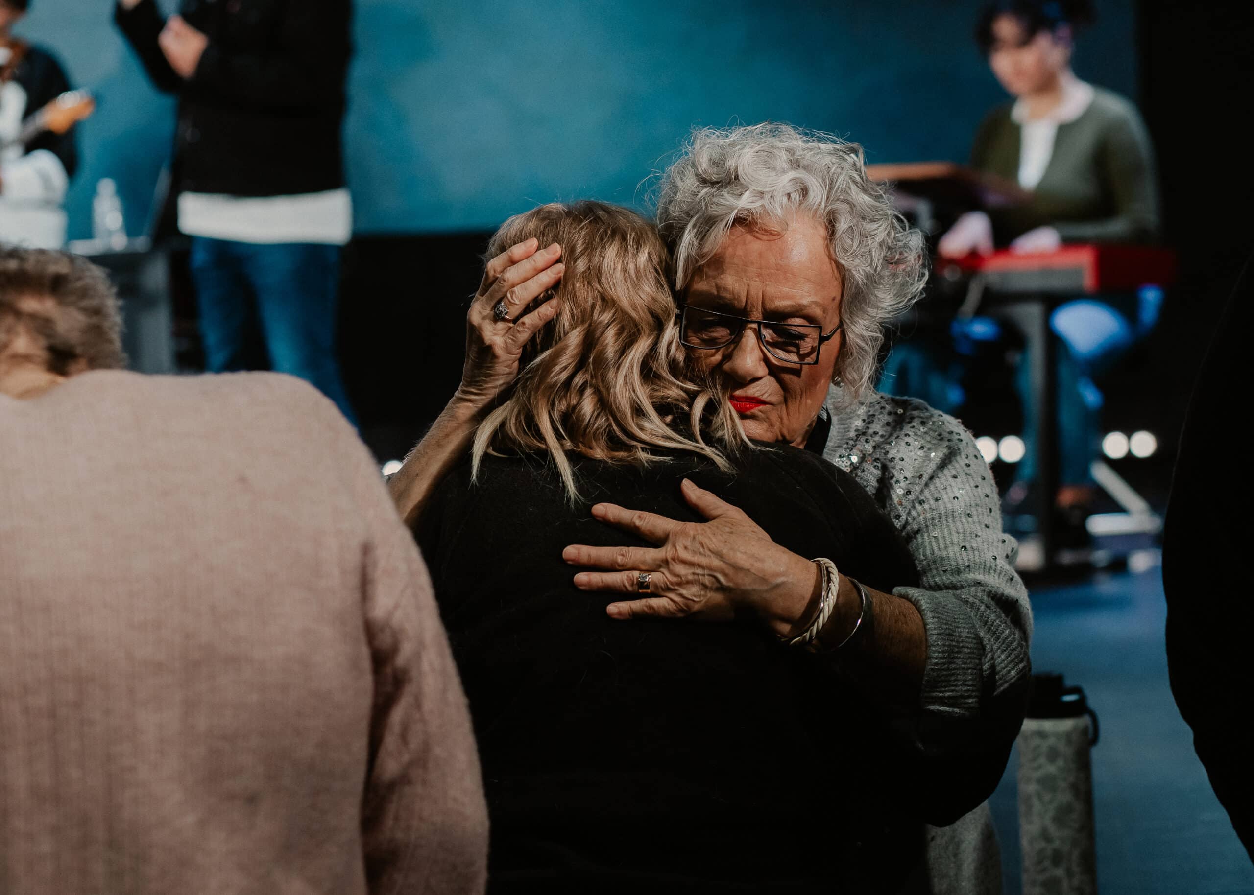 An older woman with curly gray hair and glasses embraces a younger woman with long hair. In the background, other individuals are engaged in activities, and a musician is playing an instrument. The setting appears to be a community event.