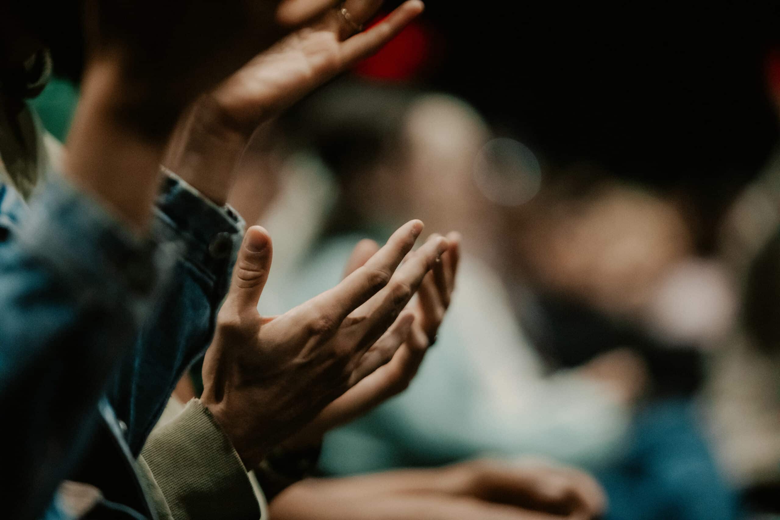 Close-up view of hands clapping, with a blurred audience in the background. The image shows multiple hands of varying skin tones, some wearing denim and others in casual attire, engaged in applause.