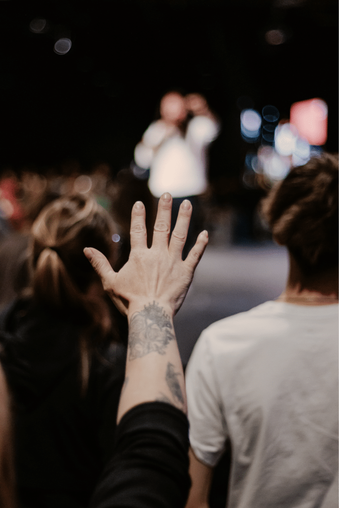 A raised hand with a tattoo is in focus with blurred figures in the background, including a person speaking on stage. The setting appears to be a large event or gathering, with mostly dark surroundings and colorful lights.
