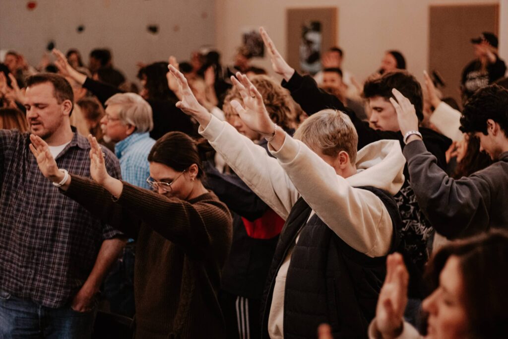 A group of people standing with their hands raised in a large indoor gathering, with rows of attendees and a softly lit room in the background.