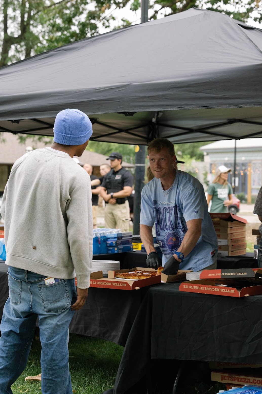 A man in a blue beanie stands at a canopy-covered table while another man hands out boxed items, with police officers and trees in the background.