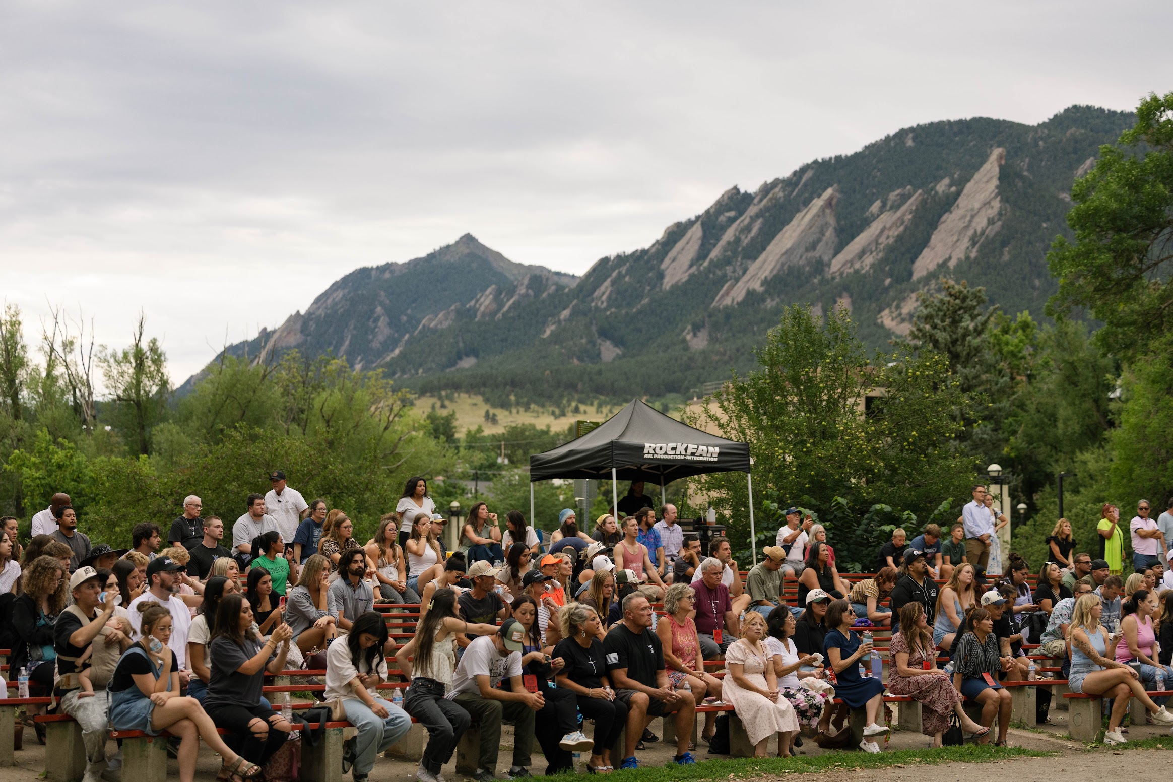 A large seated crowd attends an outdoor event in front of mountain peaks, with a black Rockeze tent and trees in the background.