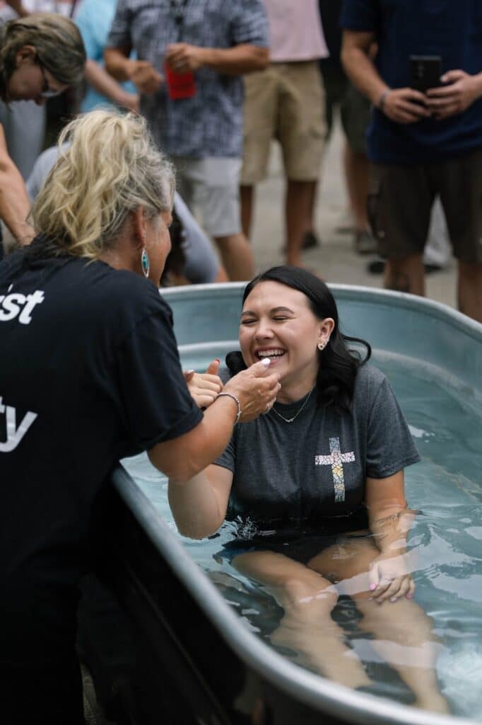 A woman in a dark T-shirt is being baptized in a metal tub outdoors, smiling as another woman stands beside her and pours water over her mouth.