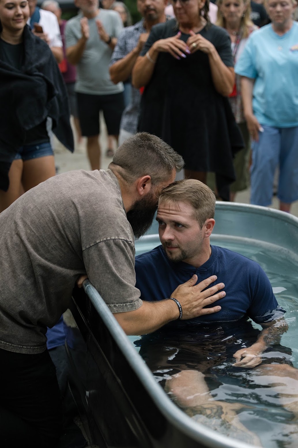 A man in a blue shirt sits in a water-filled tub while another man leans over the rim to support him, with a group of people standing behind them outdoors.