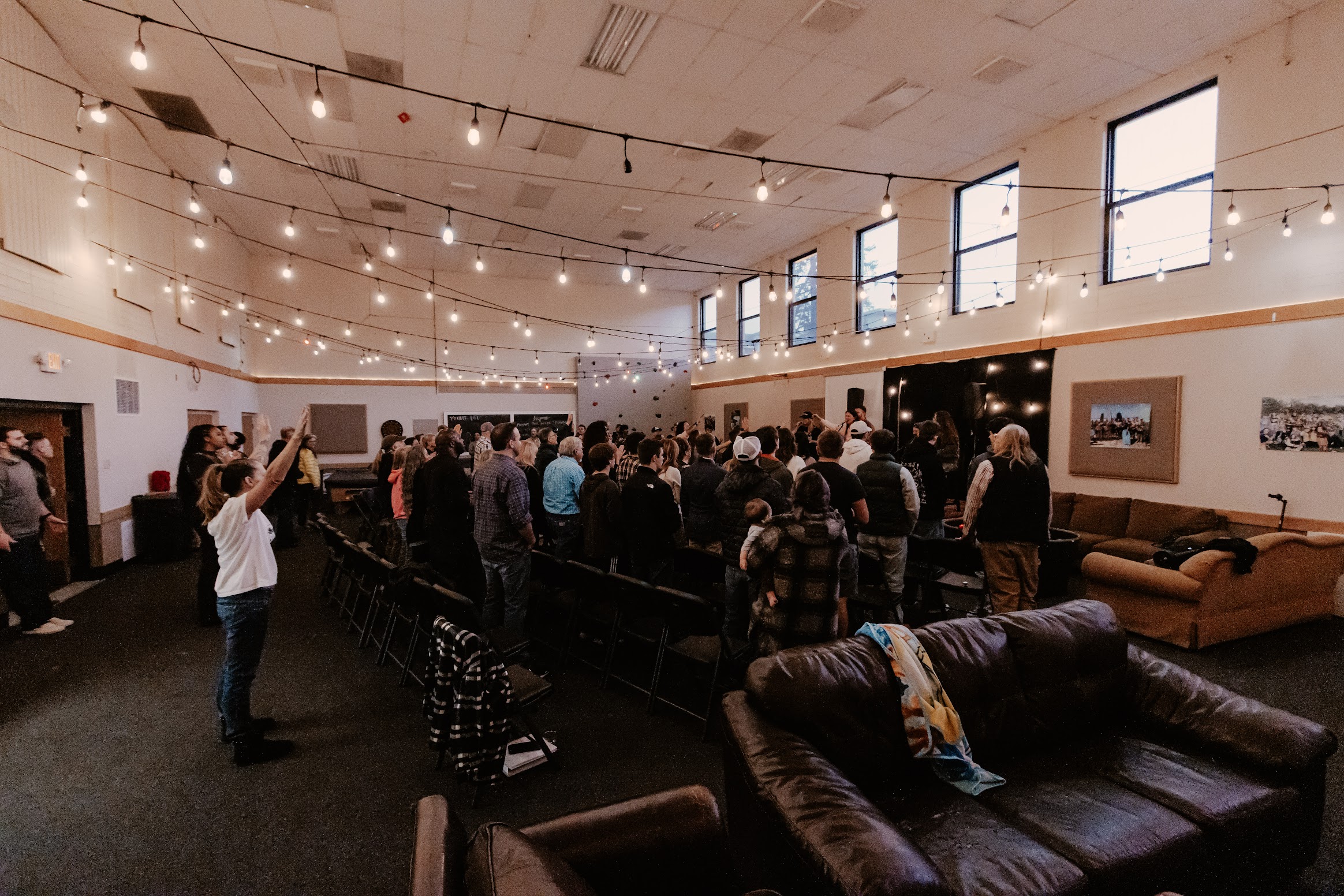 A group of people stands in a large hall with string lights and tall windows.