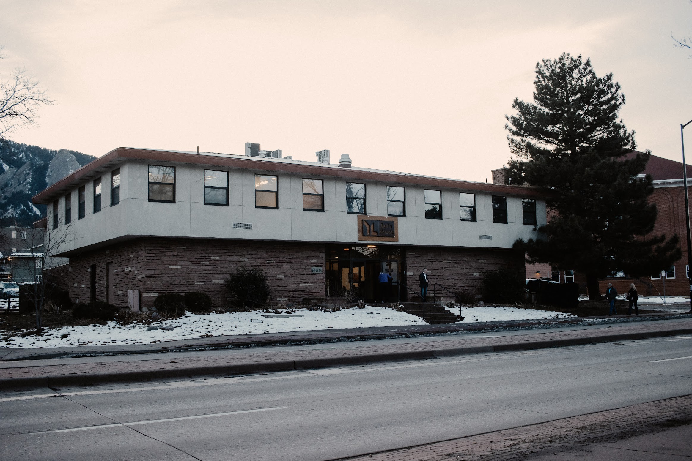 A two-story brick-and-white building with many windows stands beside a snowy sidewalk and road, with a tall evergreen tree on the right.