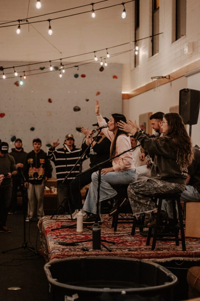 Three musicians perform seated on stools on a patterned rug in a warehouse-like venue, with string lights overhead and a small audience standing nearby.