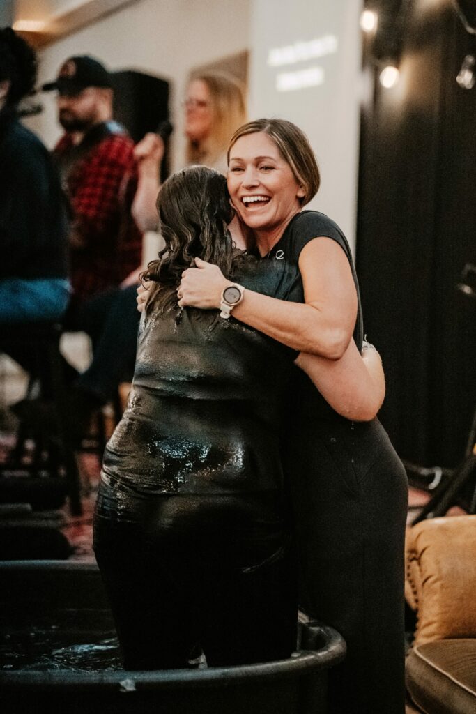 Two women hug in a room with people in the background; one wears a black dress and a wristwatch.