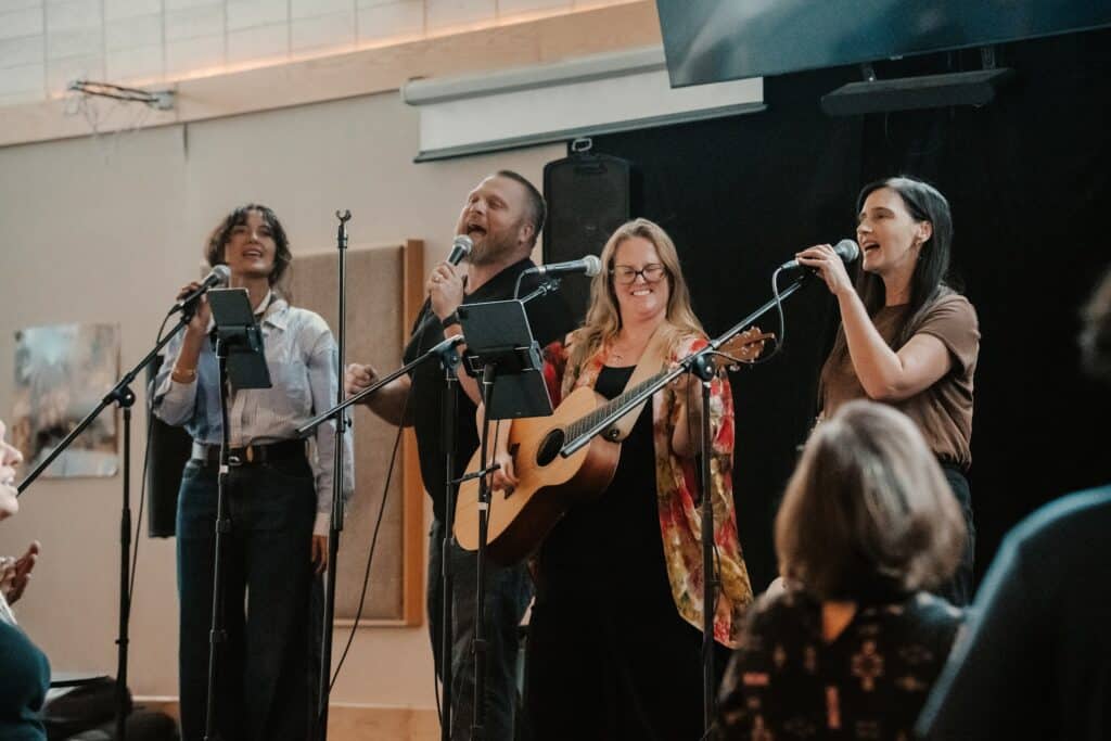 Four musicians stand at microphones performing together, with one man playing an acoustic guitar in front of a black speaker and wall panels.