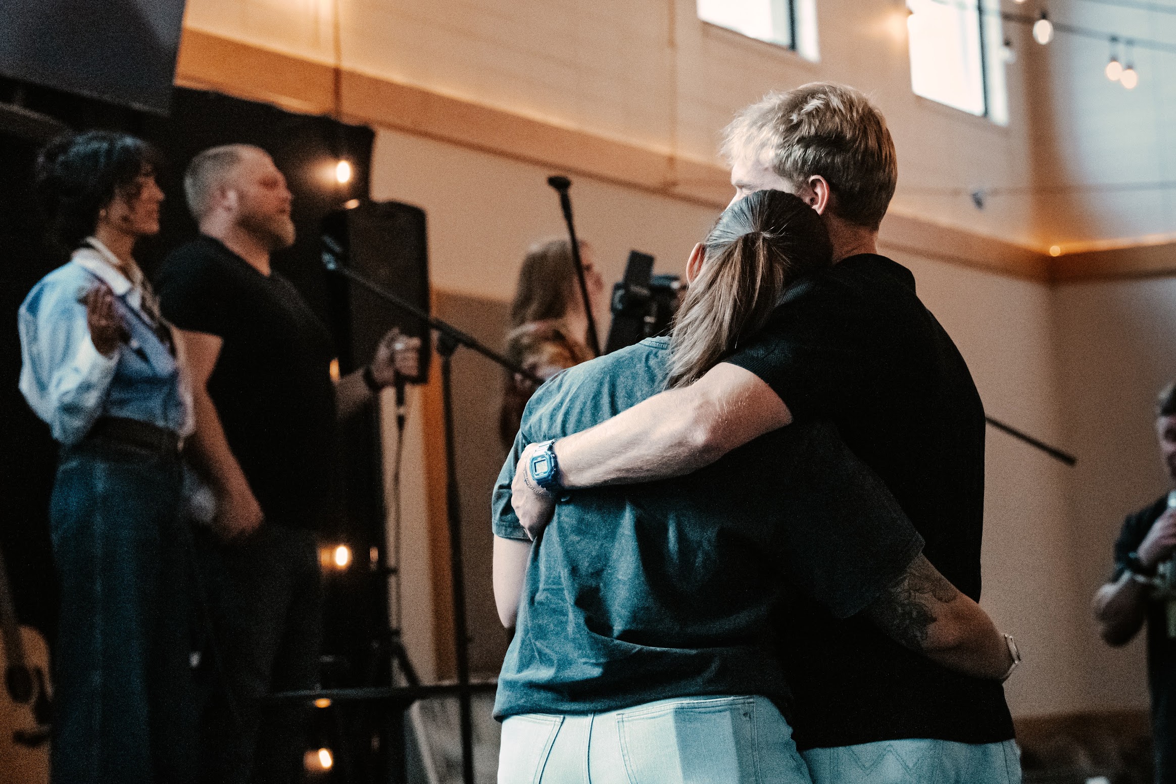 Two people embrace in a large indoor venue, with musicians and microphone stands visible in the background.